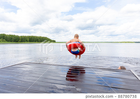 Boy jumping in a red lifebuoy from the bridge into the river, lake.A girl is hiding behind a bridge.Swim and dive.Flight, freedom, summer vacation without internet.Sky with clouds, forest on a horizon 88780738
