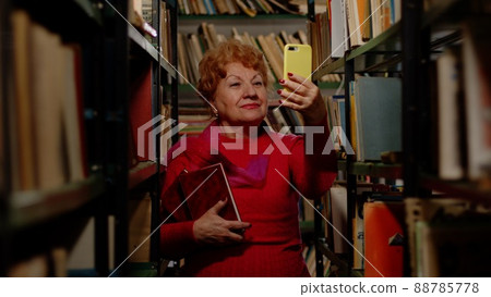 An elderly woman takes a photo with books in the library. Selfie for social networks An elderly woman takes a photo with books in the library. Selfie for social networks 88785778