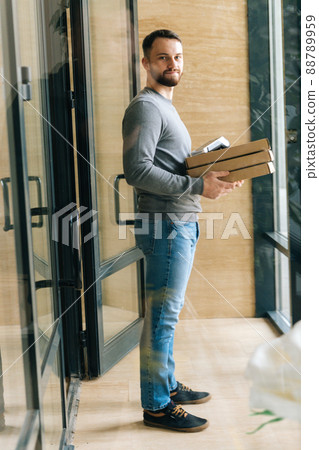 Vertical shot of courier male holding boxes pizza and payment POS terminal, standing in entrance hall of apartment or office building, by door waiting to meet customer, looking at camera. Vertical shot of courier male holding boxes pizza and payment POS terminal, standing in entrance hall of apartment or office building, by door waiting to meet customer, looking at camera. 88789959