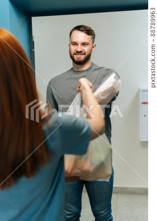Vertical shot of courier male delivering groceries bag to unrecognizable female housewife customer on doorway at home. Rear view of young woman client receiving takeout food meal from deliveryman 88789963
