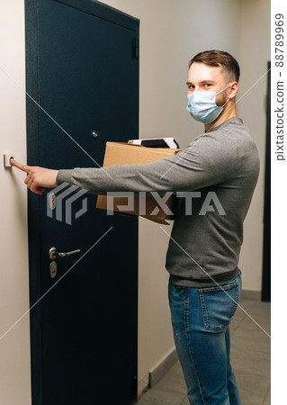 Vertical shot of handsome delivery man in medical mask ringing doorbell of customer apartment looking at camera, holding cardboard box and contactless payment POS wireless terminal for card paying. 88789969