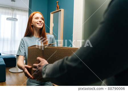 Smiling redhead young woman accepting cardboard box from unrecognizable delivery man on doorstep at apartment. Rear view of courier male delivering parcel package post to female client at home. Smiling redhead young woman accepting cardboard box from unrecognizable delivery man on doorstep at apartment. Rear view of courier male delivering parcel package post to female client at home. 88790006