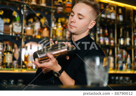 Close-up of bartender in black clothes mixing ingredients of alcoholic cocktail by shaking shaker standing behind bar counter, on blurred background of shelves with alcoholic drinks, selective focus. 88790012