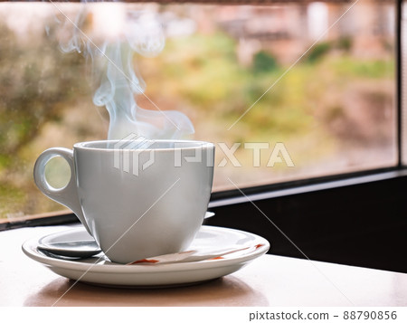 Close up of a white coffee cup on a bar table with a window in the background. Steaming cup of coffee. Close up of a white coffee cup on a bar table with a window in the background. Steaming cup of coffee. 88790856
