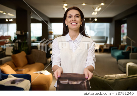 Pleasant female retail assistant, sales rep showing catalog with upholstered furniture, standing in the exposition center and smiling with cheerful toothy smile looking at camera Pleasant female retail assistant, sales rep showing catalog with upholstered furniture, standing in the exposition center and smiling with cheerful toothy smile looking at camera 88791240