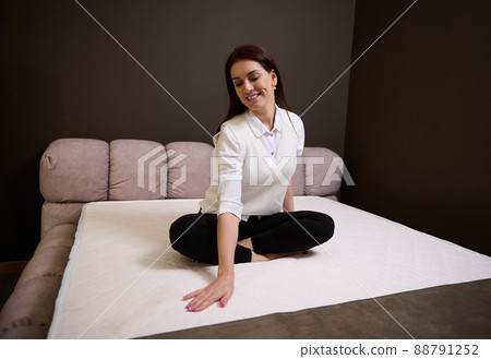 Charming woman sitting on an orthopedic mattress and touching it while shopping in furniture store exhibition center Charming woman sitting on an orthopedic mattress and touching it while shopping in furniture store exhibition center 88791252