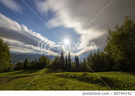 Summer night mountain panorama. Green grassy mountain clearing on cloudy evening sky copy space background, woody hills on horizon, bright dwelling lights below in valley. 88794542