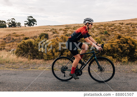 Pro bike female rider wearing helmet and sunglasses training outdoors on empty countryside road Pro bike female rider wearing helmet and sunglasses training outdoors on empty countryside road 88801507