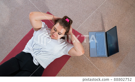 A chubby young woman is watching an online fitness lesson on a laptop. Distance sports training. A chubby young woman is watching an online fitness lesson on a laptop. Distance sports training. 88802752