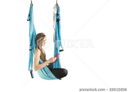 Young athletic woman hang in hammock during an aerial gymnastics or fly yoga workout has break and holding phone. Young athletic woman hang in hammock during an aerial gymnastics or fly yoga workout has break and holding phone. 88810806