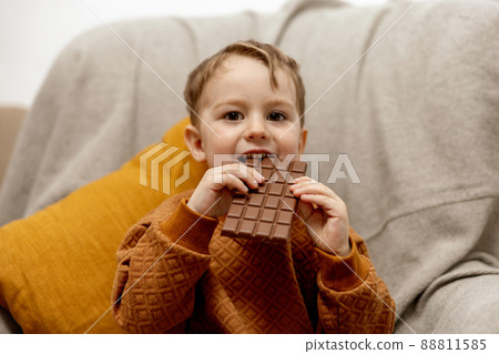 Little adorable boy sitting on the couch at home and eating chocolate bar. Child and sweets, sugar confectionery. Kid enjoy a delicious dessert. Preschool child with casual clothing. Positive emotion. 88811585