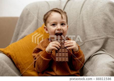 Little adorable boy sitting on the couch at home and eating chocolate bar. Child and sweets, sugar confectionery. Kid enjoy a delicious dessert. Preschool child with casual clothing. Positive emotion. 88811586