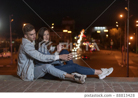Young caucasian couple is sitting on the pavement hugging each other against the backdrop of the lights of the night city. Date and relationship. Side view Young caucasian couple is sitting on the pavement hugging each other against the backdrop of the lights of the night city. Date and relationship. Side view 88813428