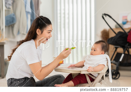 Side view mother feeding her asian baby daughter with pumpkin mashed or vegetable mash on rubber spoon.Mom trying to feed little baby boy at home enjoy and spending time together.Baby feeding Concept 88819757