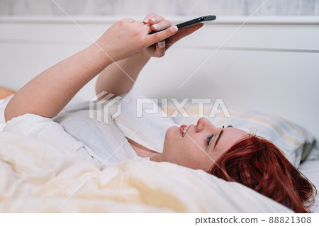 young woman smiling, taking a selfie with cell phone, lying on her bedroom bed. young woman smiling, taking a selfie with cell phone, lying on her bedroom bed. 88821308