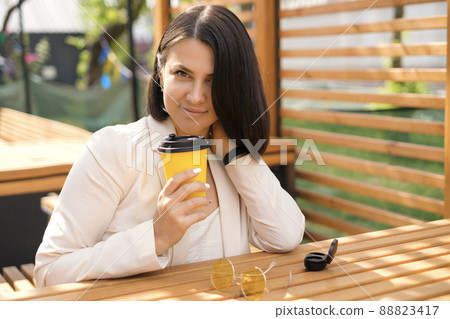 A young beautiful brunette woman is sitting at a table in the courtyard of a restaurant and drinking coffee during a break from a disposable cup 88823417