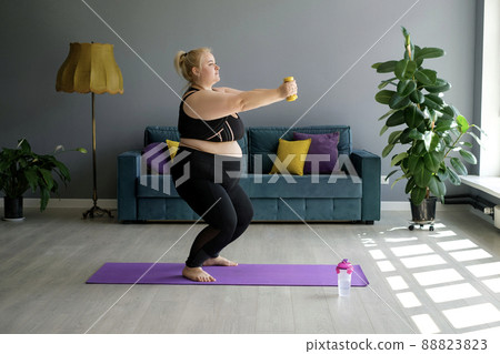 Young woman trains with yellow dumbbells and does squats at the same time. Concept of doing sports at home. Overweight lady training at home in front of sofa in room . Plus size strengthens biceps Young woman trains with yellow dumbbells and does squats at the same time. Concept of doing sports at home. Overweight lady training at home in front of sofa in room . Plus size strengthens biceps 88823823