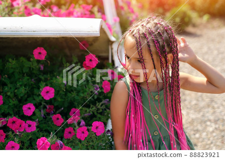 A beautiful cute girl with pink Afro-pigtails from kanekalon stands in front of a flower bed with flowers. a warm summer day. Petunia flowers with a pronounced sweet smell. zizi pigtails. 88823921