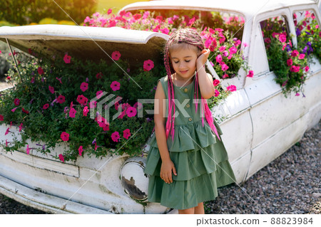 A girl with fashionable zizi pigtails from kanekalon closed her eyes from enjoying the smell of flowers. A kid is standing by a decorative car used instead of a flowerbed. Landscape Design Ideas 88823984