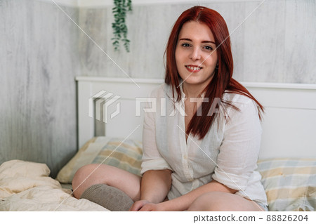 smiling young woman, sitting on her bedroom bed, looking at camera. 88826204