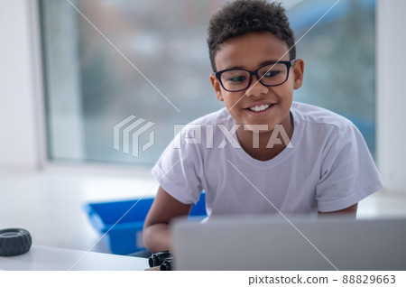 A smiling dark-skinned boy sitting at the laptop A smiling dark-skinned boy sitting at the laptop 88829663