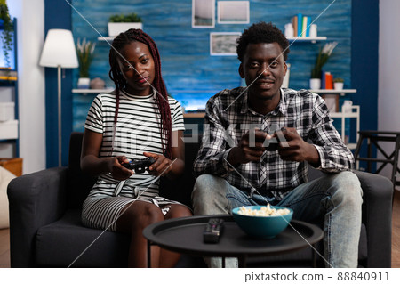 Relaxed african american couple playing video games on console while having a good time sitting at home on the sofa. Joyful people enjoying free time while having fun together in living room. 88840911