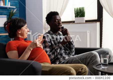 Pregnant caucasian woman laughing at african american man singing at remote control while sitting on sofa in living room. Joyful adult couple acting funny while expecting baby at home. 88841100