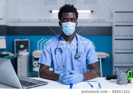 Hospital medical staff sitting at table in clinic cabinet while wearing surgical gloves and virus protection mask. Healthcare nurse sitting at office desk while wearing facemask. 88841102