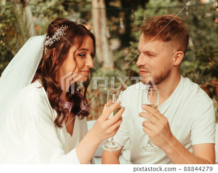 Stylish, smiling groom and cute blonde bride with curly hair in a white dress are sitting on the rug with glasses of champagne. grain effect Stylish, smiling groom and cute blonde bride with curly hair in a white dress are sitting on the rug with glasses of champagne. grain effect 88844279