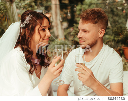 Stylish, smiling groom and cute blonde bride with curly hair in a white dress are sitting on the rug with glasses of champagne. grain effect Stylish, smiling groom and cute blonde bride with curly hair in a white dress are sitting on the rug with glasses of champagne. grain effect 88844280