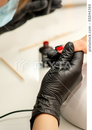 Manicure varnish painting. Close-up of a manicure master wearing rubber black gloves applying red varnish on a female fingernail in the beauty salon Manicure varnish painting. Close-up of a manicure master wearing rubber black gloves applying red varnish on a female fingernail in the beauty salon 88844369