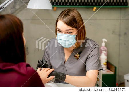 Portrait of young caucasian manicure master wearing protective mask painting female nails in a beauty salon Portrait of young caucasian manicure master wearing protective mask painting female nails in a beauty salon 88844371