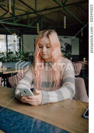 Young curious woman with pink hair likes millennial beautiful girl looking with interest watching smiling lady sitting nearby in cafe using smartphone Young curious woman with pink hair likes millennial beautiful girl looking with interest watching smiling lady sitting nearby in cafe using smartphone 88844805