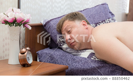 An adult man 40-45 years old is fast asleep in his bed without hearing a loud alarm clock 88850462