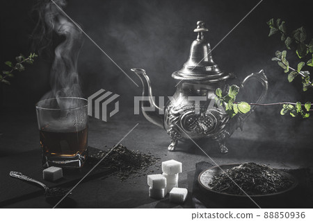 Traditional Moroccan teapot, with a steaming cup of tea, sugar and mint, in a smoky black atmosphere. Moroccan tea concept. Traditional Moroccan teapot, with a steaming cup of tea, sugar and mint, in a smoky black atmosphere. Moroccan tea concept. 88850936