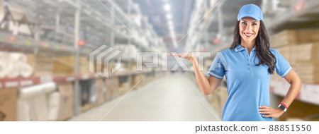 Young delivery woman in work clothes in front of a huge warehouse her left hand offers free space for information 88851550