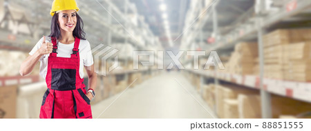 A warehouse worker points her finger up. Smiling young woman in work clothes in front of a huge warehouse 88851555