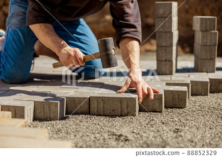 Young man laying gray concrete paving slabs in house courtyard on gravel foundation base. Master lays paving stones. Garden brick pathway paving by professional paver worker. Repairing sidewalk. Young man laying gray concrete paving slabs in house courtyard on gravel foundation base. Master lays paving stones. Garden brick pathway paving by professional paver worker. Repairing sidewalk. 88852329