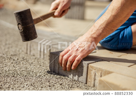 Young man laying gray concrete paving slabs in house courtyard on gravel foundation base. Master lays paving stones. Garden brick pathway paving by professional paver worker. Repairing sidewalk. Young man laying gray concrete paving slabs in house courtyard on gravel foundation base. Master lays paving stones. Garden brick pathway paving by professional paver worker. Repairing sidewalk. 88852330