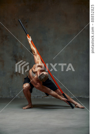 A fighter shirtless in black shorts, stands in a fighting artificially pose and holds a fight stick on studio background. A fighter shirtless in black shorts, stands in a fighting artificially pose and holds a fight stick on studio background. 88852825