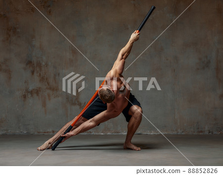 A fighter shirtless in black shorts, stands in a fighting artificially pose and holds a fight stick on studio background. 88852826