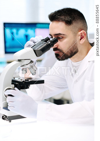 Young bearded man in lab coat and gloves looking at bacteria in microscope Young bearded man in lab coat and gloves looking at bacteria in microscope 88855645