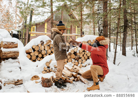 Young father sitting on squats and passing firewood to his son against woodpile Young father sitting on squats and passing firewood to his son against woodpile 88855698