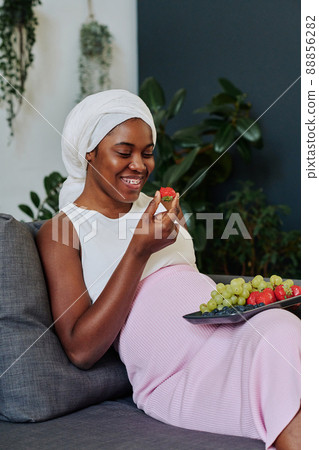 African muslim woman in hijab sitting on sofa and eating fresh fruits during her pregnancy African muslim woman in hijab sitting on sofa and eating fresh fruits during her pregnancy 88856282