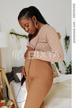 African pregnant woman using her mobile phone for enjoying music while standing in bedroom African pregnant woman using her mobile phone for enjoying music while standing in bedroom 88856408