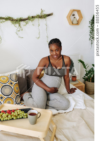 Portrait of African pregnant girl smiling at camera sitting on bed with fruits and tea on tray Portrait of African pregnant girl smiling at camera sitting on bed with fruits and tea on tray 88856455