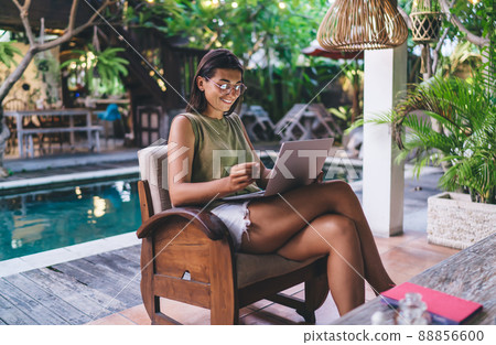 Happy ethnic woman using laptop in cafe near pool during summer holidays 88856600