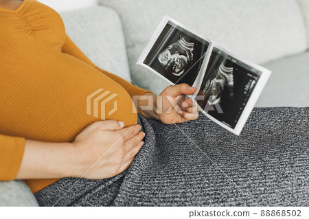 Pregnant woman looking at first ultrasound photo of her baby, caressing her belly happily awaiting the birth of her child. First trimester pregnancy Pregnant woman looking at first ultrasound photo of her baby, caressing her belly happily awaiting the birth of her child. First trimester pregnancy 88868502