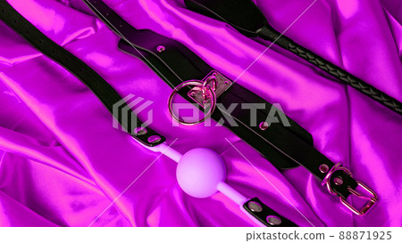 Adult sex games. BDSM items. Leather collar, pink gag ball and whip on a red satin sheet. 88871925