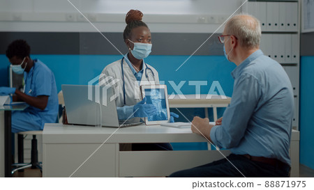 Young medic showing modern tablet with x ray scan to senior ill man at healthcare facility with plexiglass wall. Doctor holding radiography result on digital device for professional checkup 88871975